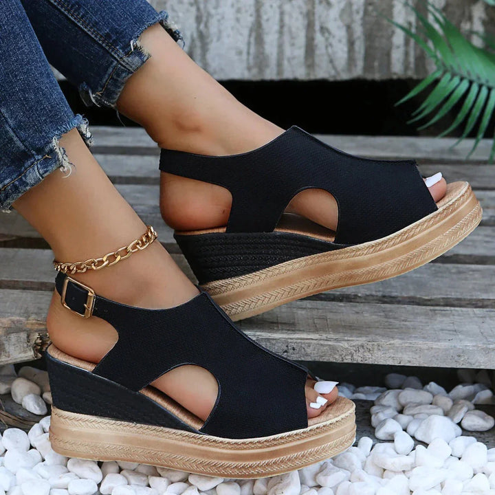 Black wedge sandals with wide strap, peep-toe cutouts, and gold chain ankle strap