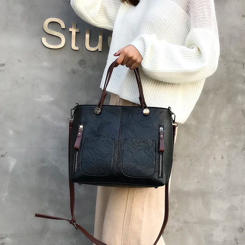 Black PU leather crossbody tote with twin front pockets, top handles, and long shoulder strap.