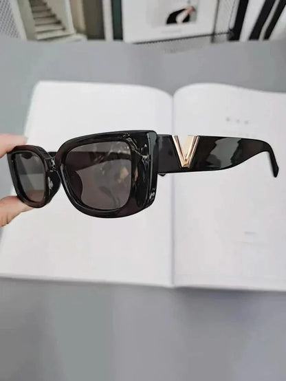 Black sunglasses held in hand, dark lenses, gold V temple accent.