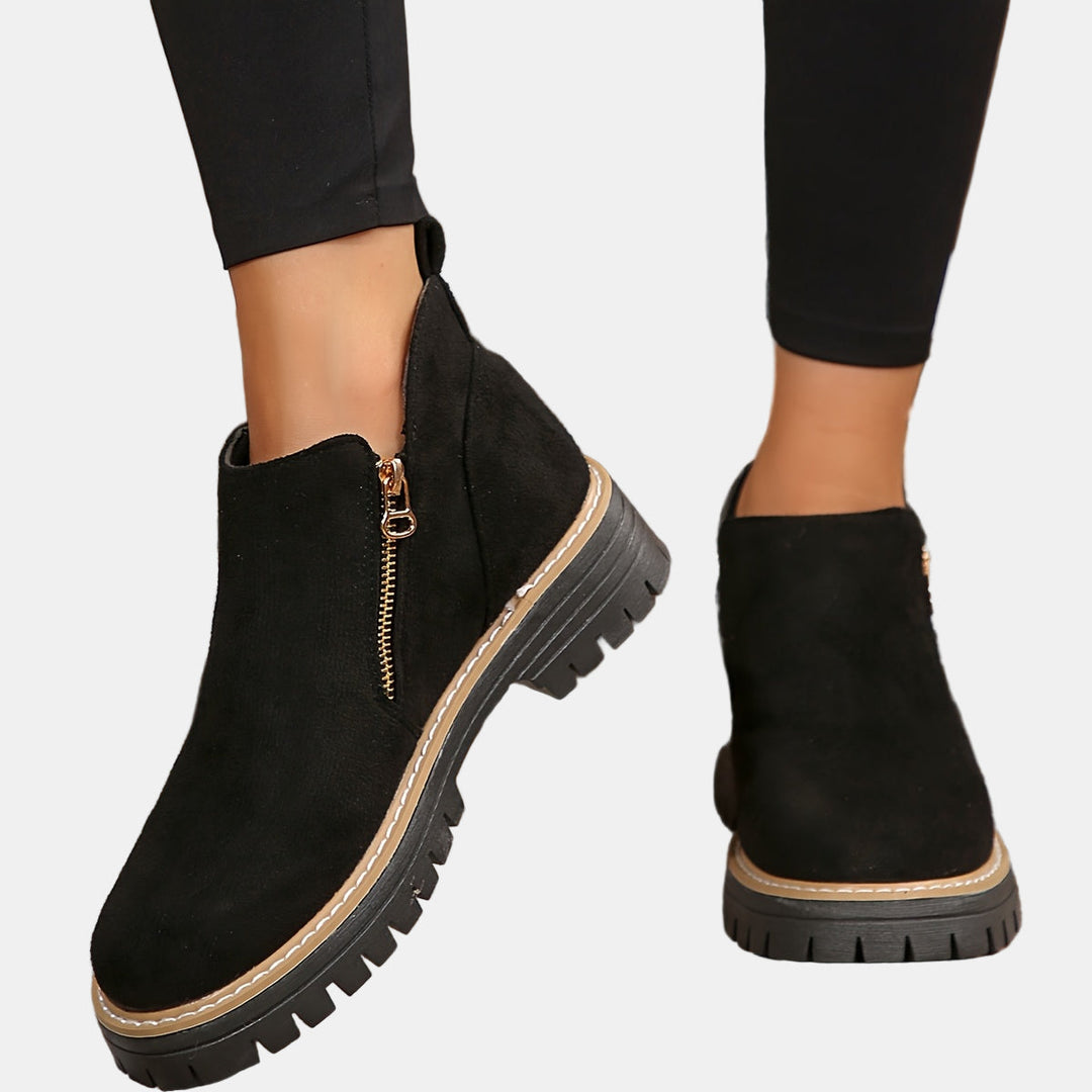 Black suede ankle boots with side zipper, tan stitching, and chunky lug sole.