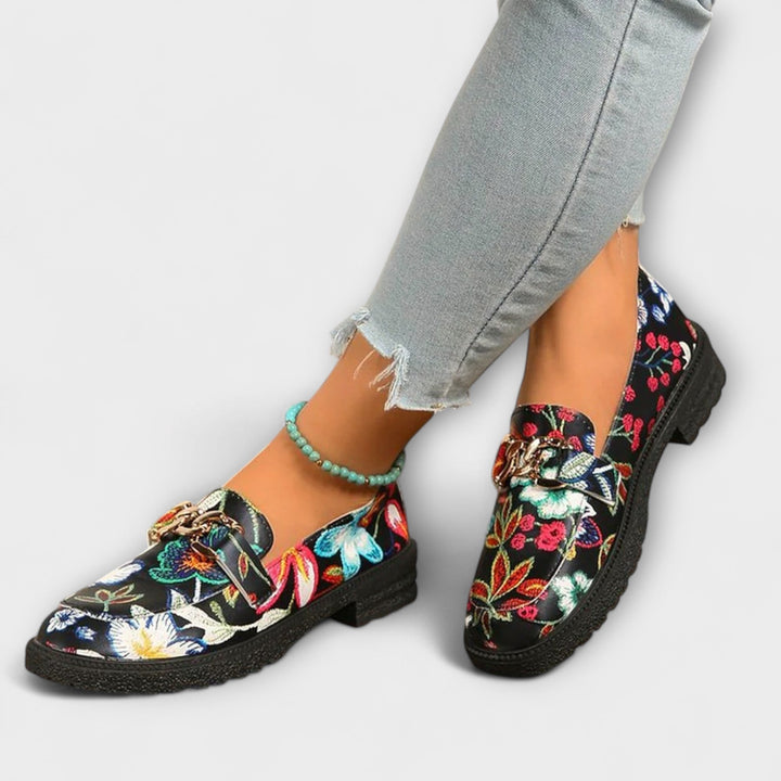 Multicolour floral loafers on feet, black slip-on style with gold chain buckle and chunky sole